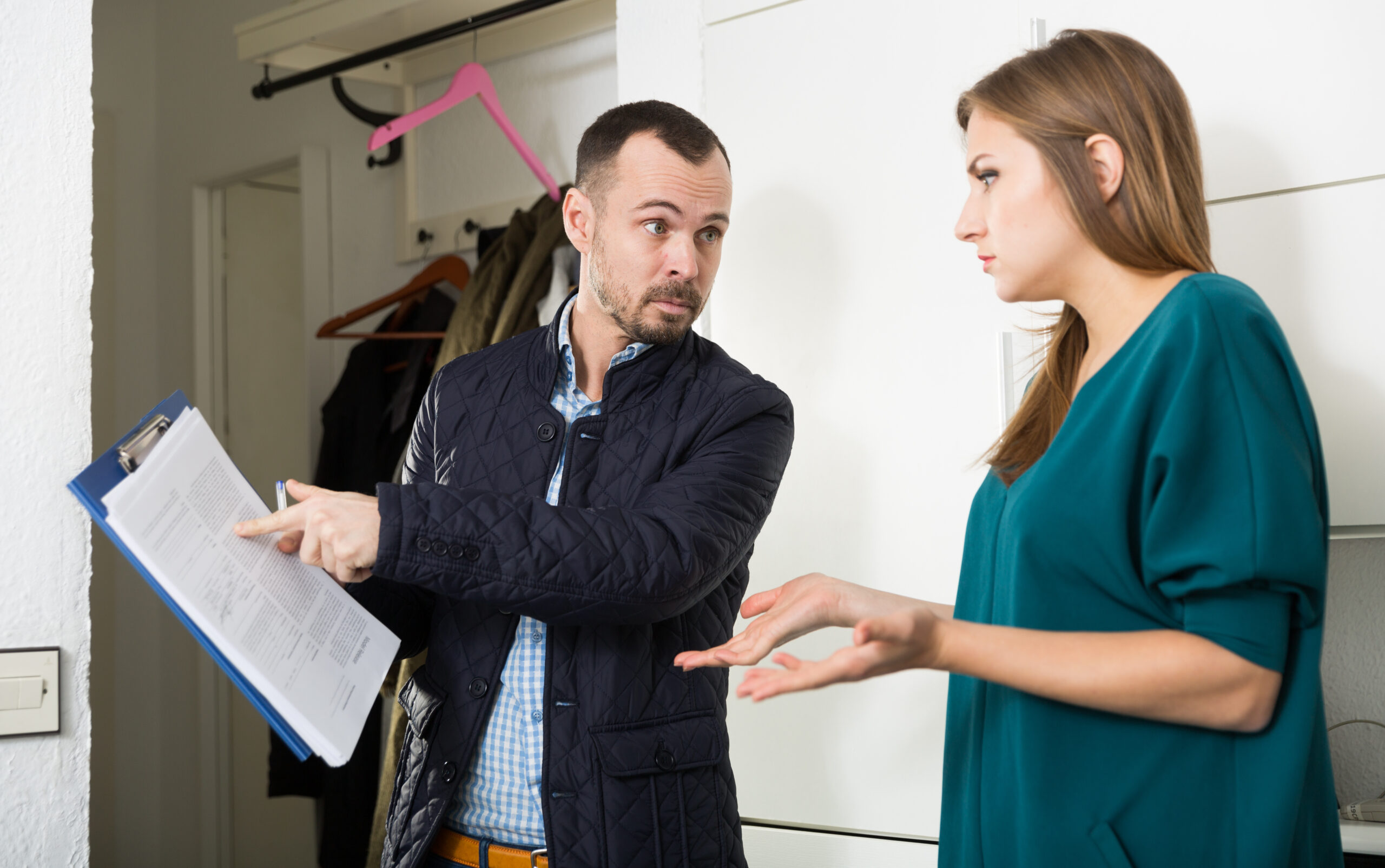 A frustrated, professionally dressed man angrily pointing at his clipboard as he confronts a confused woman in her home.