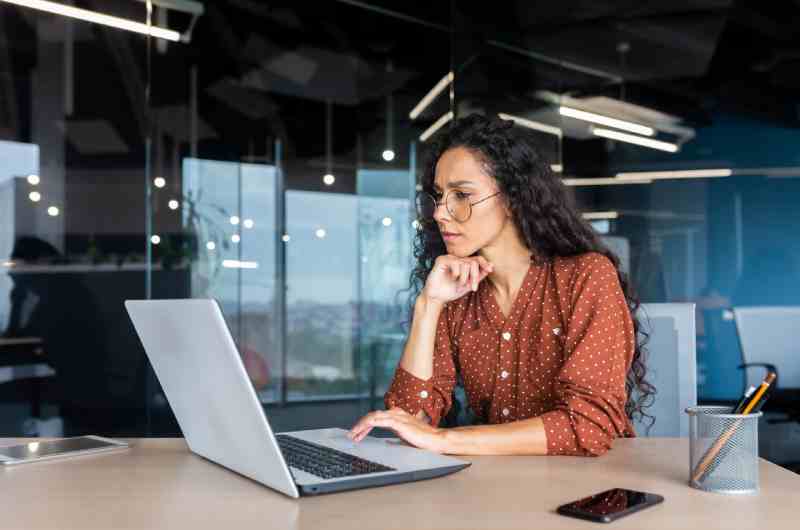 A woman with large glasses and a polka-dot blouse sitting at her desk while staring contemplatively at her computer.