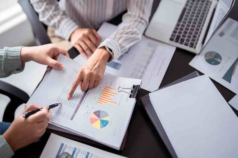 A shot of the hands of two business people sitting at a desk pointing at papers with important data on them.