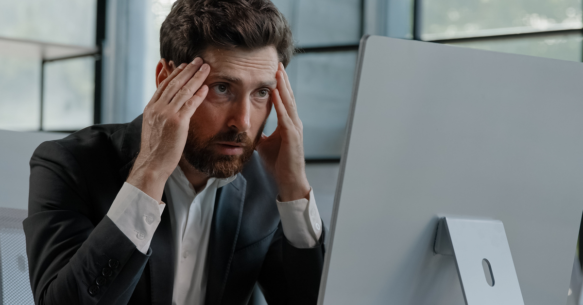 An older man wearing a suit, sitting at his computer desk staring in frustration at the computer screen as he rubs his temples.