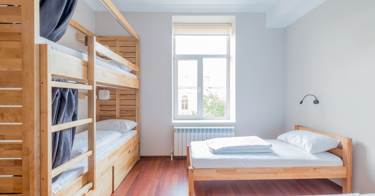 A white-walled dorm room, unfurnished aside from the two bare beds up against the wall and the bunk bed against the opposite wall.