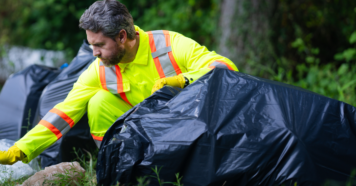 A man in high visibility gear kneeling on the ground as he stuffs multiple large trash bags with various items.