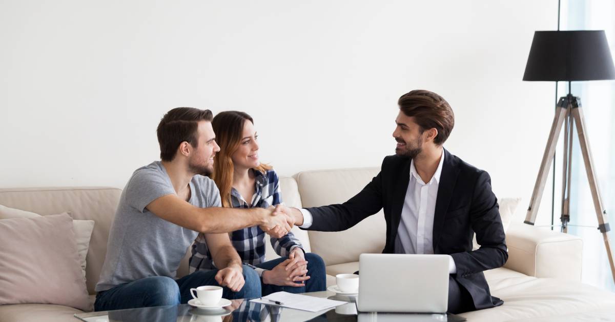 A man in a suit sitting next to a couple on a couch. His laptop is in front of him, and he shakes the other man's hand.