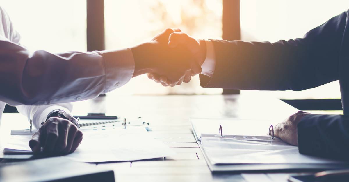 Two professionally dressed, nondescript workers reaching across a work desk to shake each other's hand.