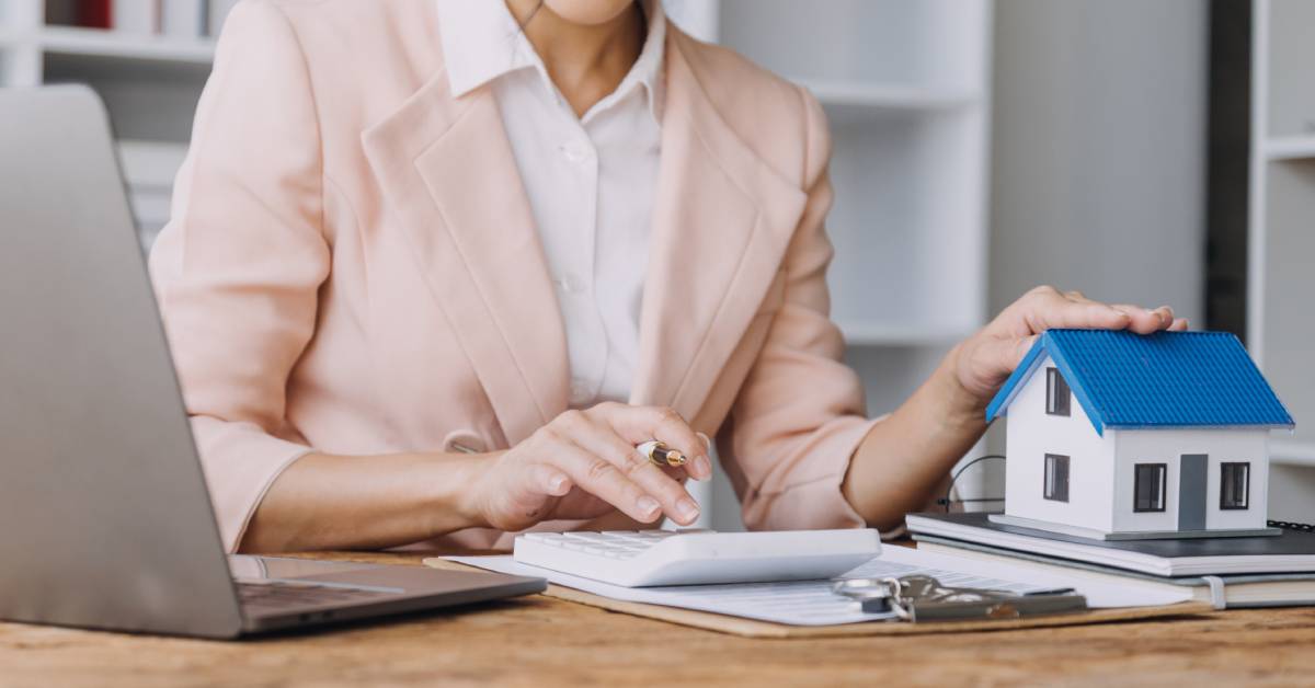 A real estate agent in a pink suit sitting at her computer desk while she types on a calculator and holds a miniature model home.