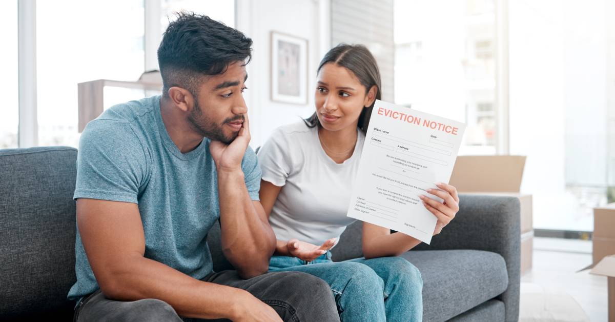 A disappointed couple sitting next to each other on their gray couch while the female partner holds up their eviction notice.