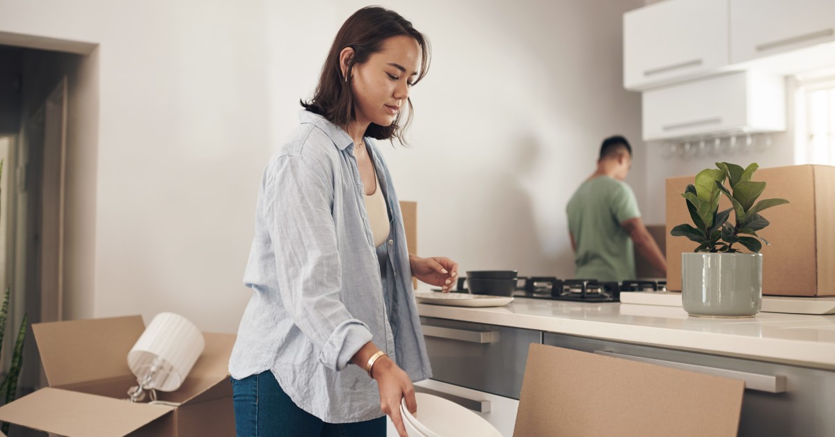 A young woman removing white plates from a brown moving box. A man is in the background along with other moving boxes.