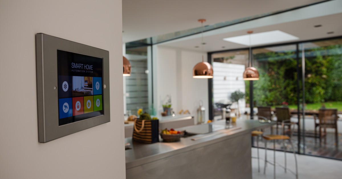 A wall-mounted touch screen that reads, "smart home" with multi-colored buttons. There is a kitchen in the background.