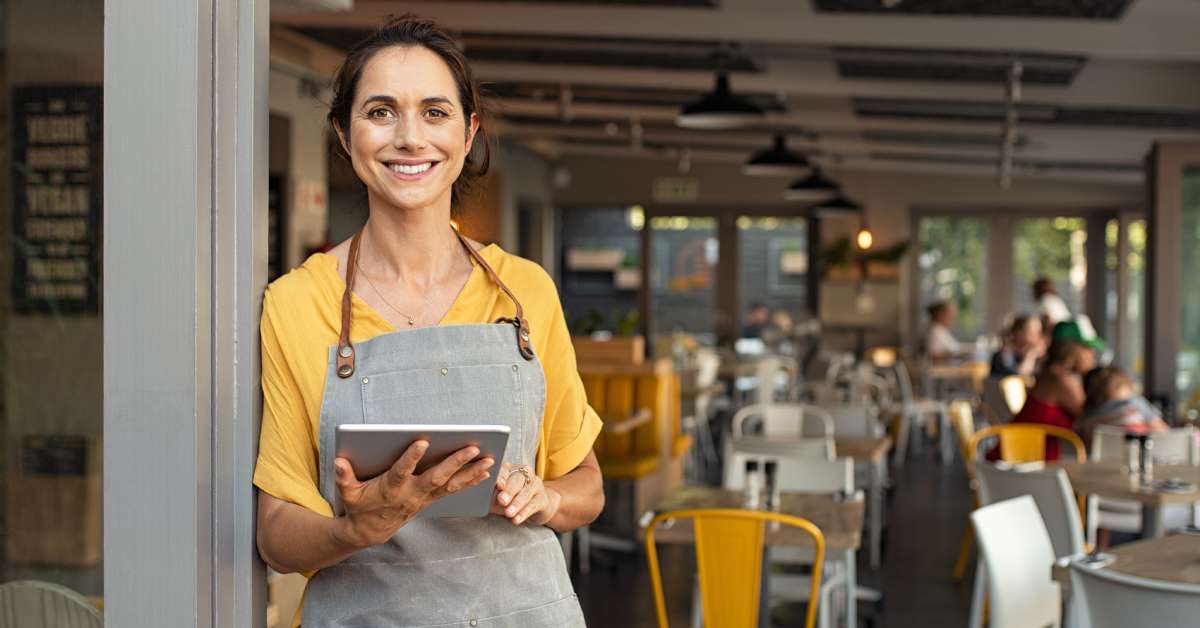 A woman in a yellow t-shirt and grey apron is smiling at the camera as she holds an electronic tablet and leans against a pole.