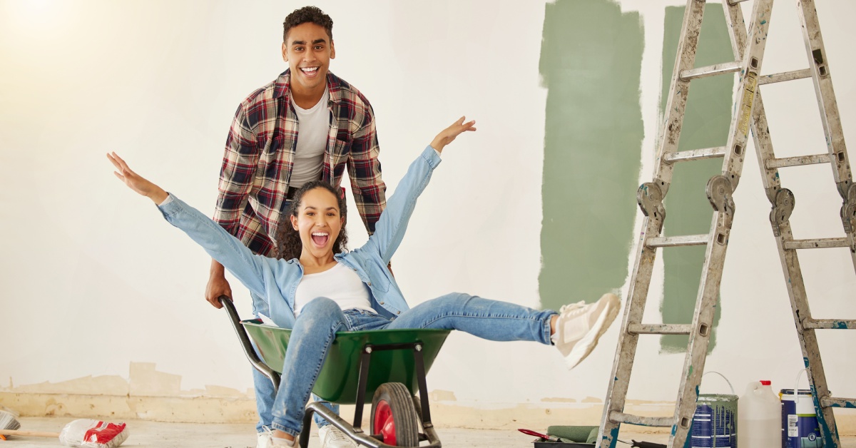 A man pushes a happy woman in a green wheelbarrow from behind. They are next to a ladder an a wall that is not fully painted.