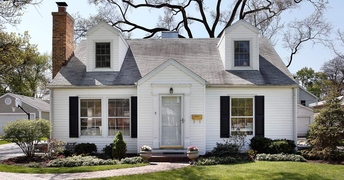 A white suburban home with black shutters and a red brick chimney. A brick path cuts to the left through their lush front lawn.