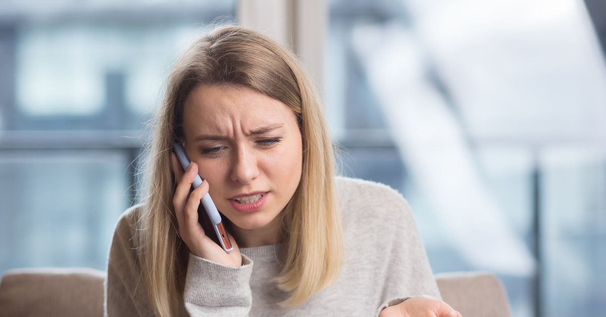 A woman wearing a grey sweatshirt sitting on her couch is on a phone call feeling confused and frustrated.