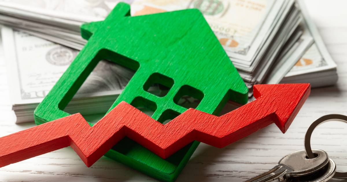 A green house sits on a stack of hundred dollar bills with a red arrow trending upward next to a set of house keys.