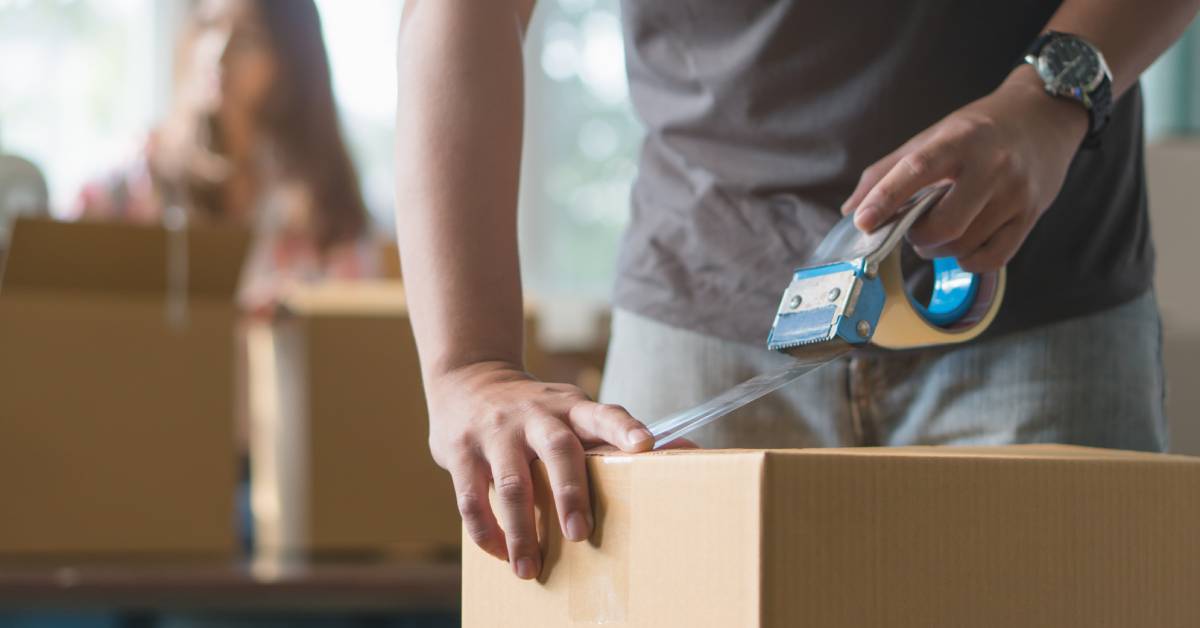 A man using clear packing tape to secure a cardboard box for moving with other boxes in the blurred background.