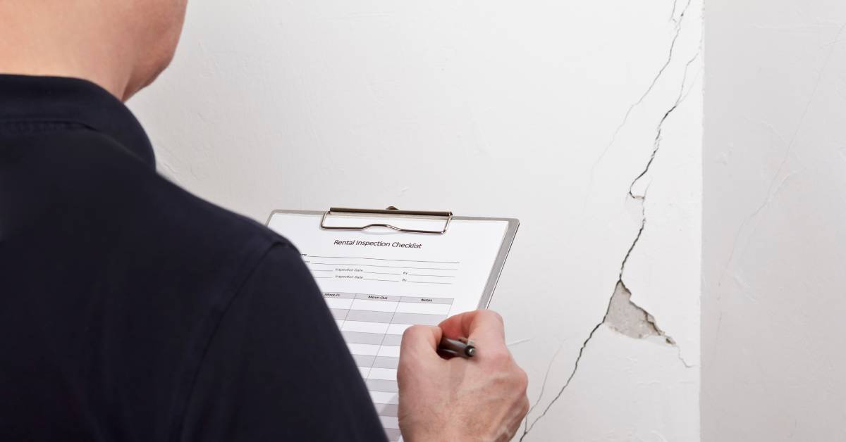 A man holding a clipboard with a rental inspection checklist takes note of a large crack on a white wall.